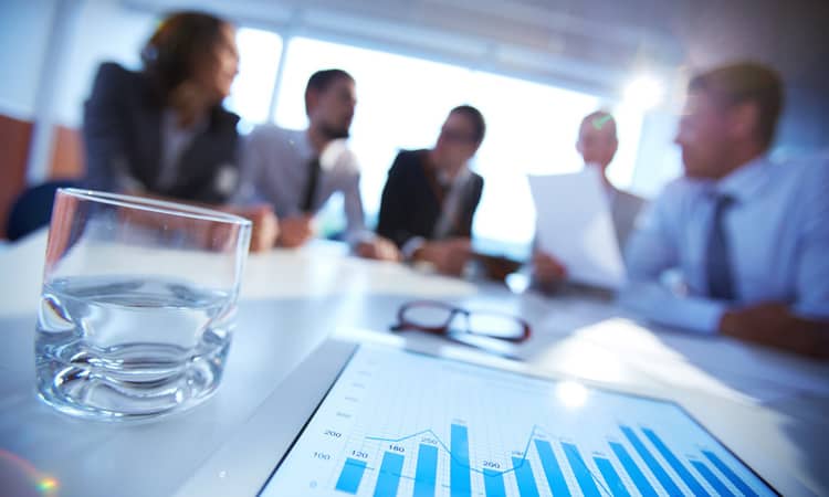 A half full glass of water with business people at a table in the background.